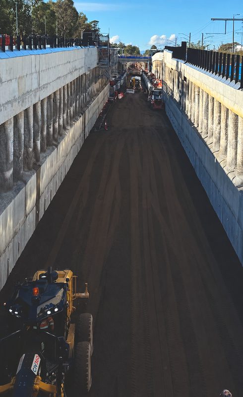 CIV360 grader at the end of a long lowered-rail trench lined with piled concrete walls