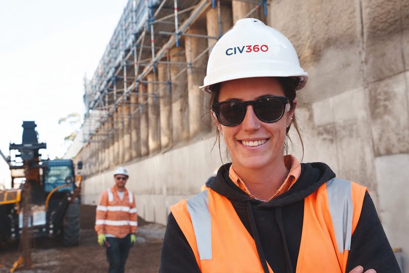 Smiling CIV360 site engineer in hard hat and sunglasses beside a piled retaining wall with scaffolding