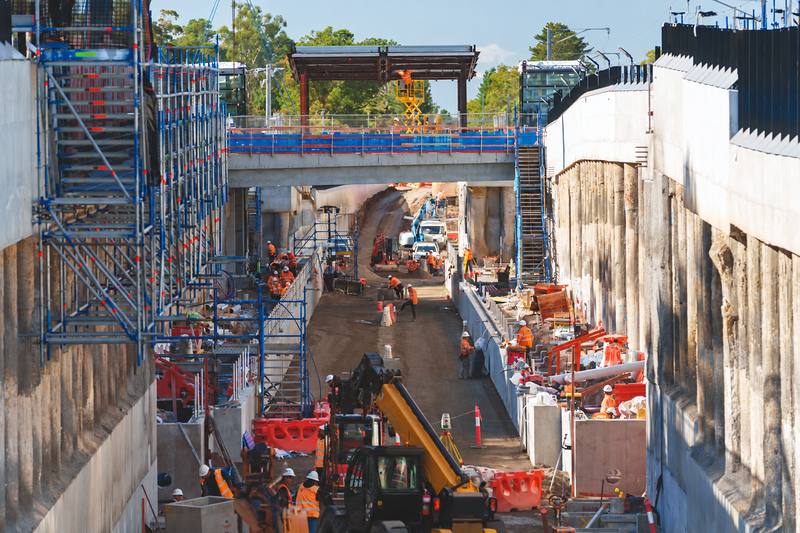 Busy lowered-rail trench with workers, a telehandler and a new pedestrian bridge under construction