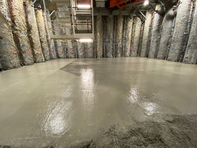 Freshly poured concrete base slab at the bottom of the shaft, with mesh-covered pile walls and a stair tower behind