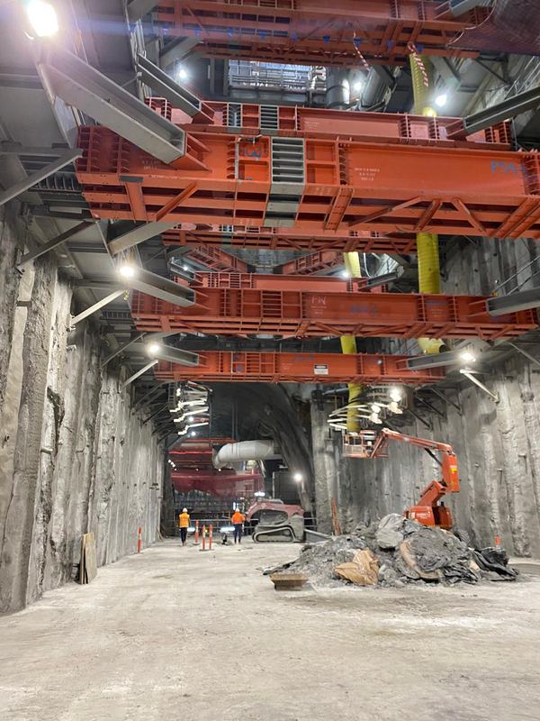 Two workers walking across the excavated floor beneath red steel props towards a tunnel opening