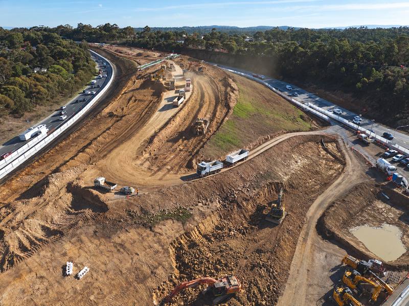 Aerial view of bulk earthworks beside a freeway, with excavators loading trucks and a dozer shaping the batter