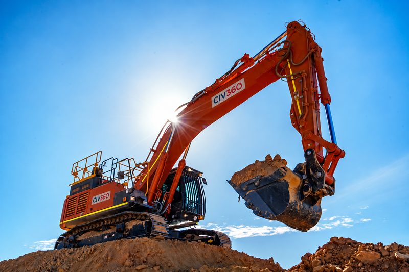 Low-angle view of a CIV360 excavator raising a full bucket against a sunlit blue sky