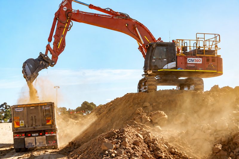 Excavator on a spoil mound loading a truck and dog trailer in a cloud of dust