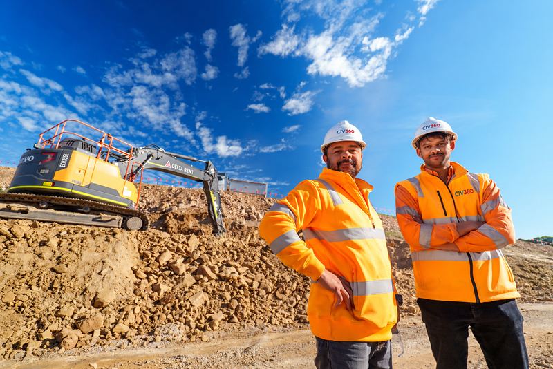 Two CIV360 crew members in hi-vis and hard hats in front of an excavator on a rock earthworks site