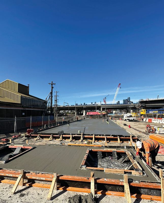 Concrete finisher working on a freshly poured slab at a rail yard, with locomotives and the city skyline behind
