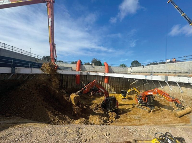 Three excavators working the floor of the shaft as a long-reach grab lifts spoil out, under a blue sky