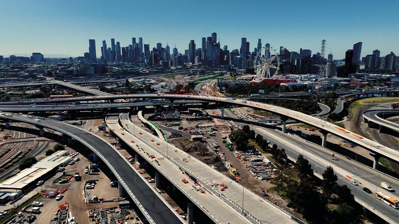 Aerial view of elevated freeway ramps under construction beside a rail yard, with the Melbourne skyline behind